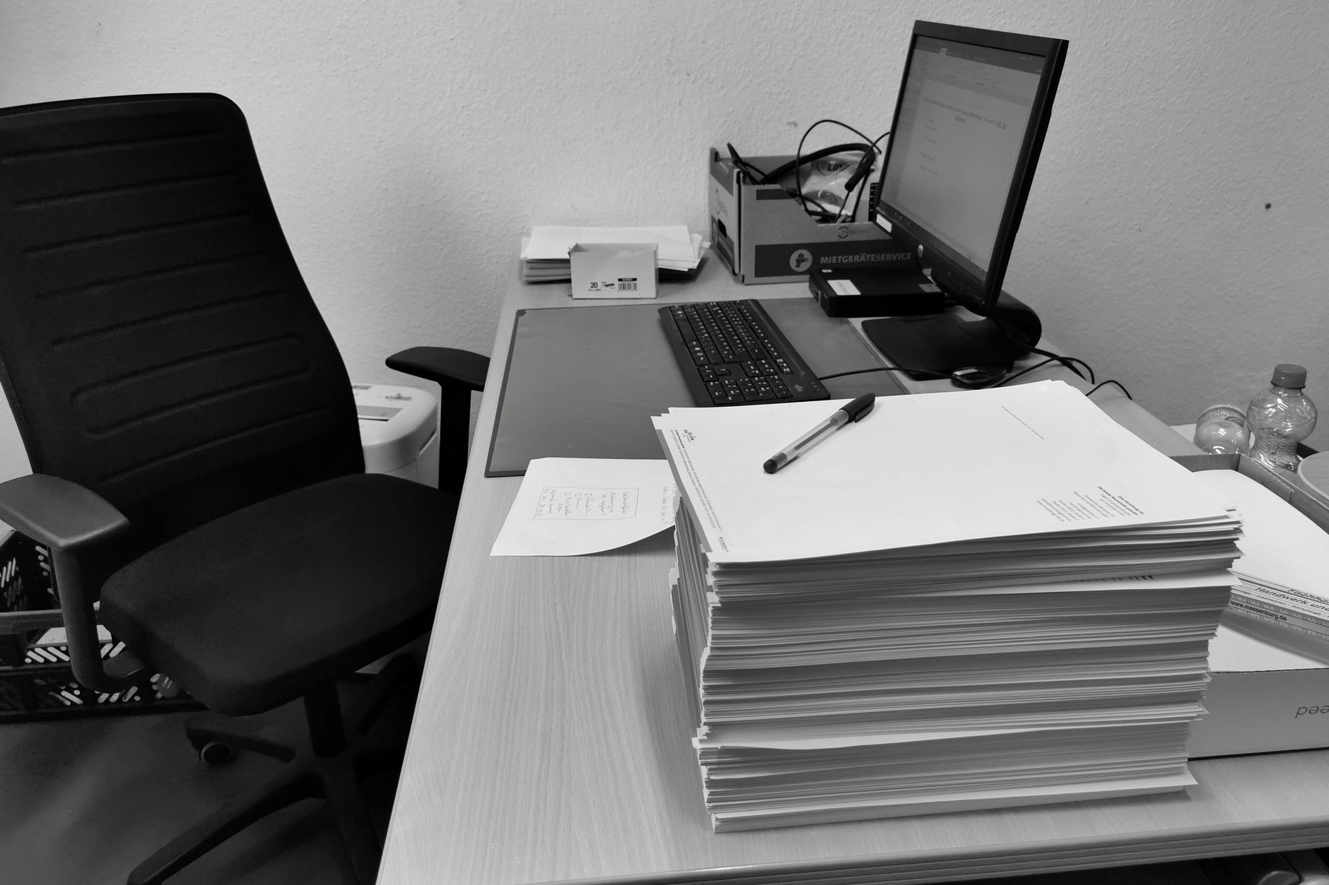 Schwarzweißbild eines überfüllten Büroschreibtischs mit Computer, Stuhl und Papierkram.